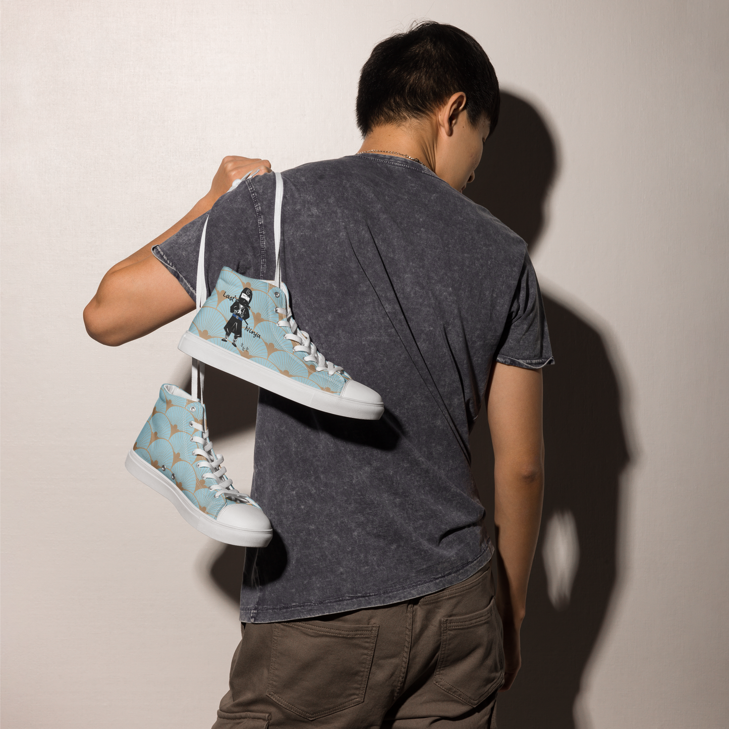 Person holding a pair of blue high-top sneakers with a playful ninja graphic design and white laces, featuring a text 'Last ninja' against a plain background
