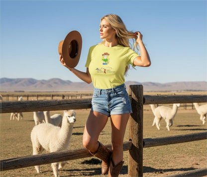 Woman in a yellow t-shirt with a cowgirl graphic and denim shorts standing in front of a wooden fence with alpacas in the background.