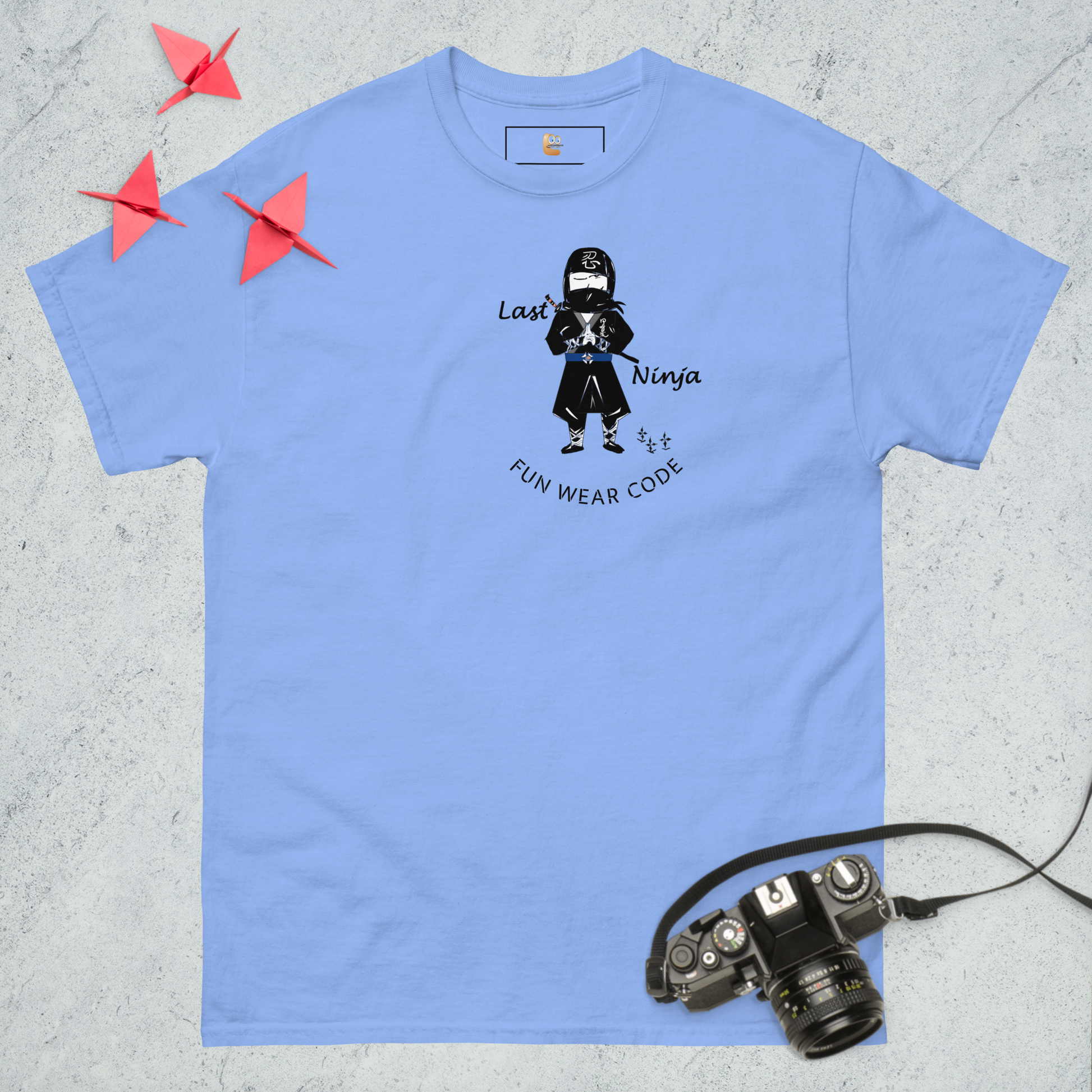 Blue t-shirt with a playful ninja graphic design and text "FUN WEAR CODE", placed on a textured surface with a camera and origami birds.