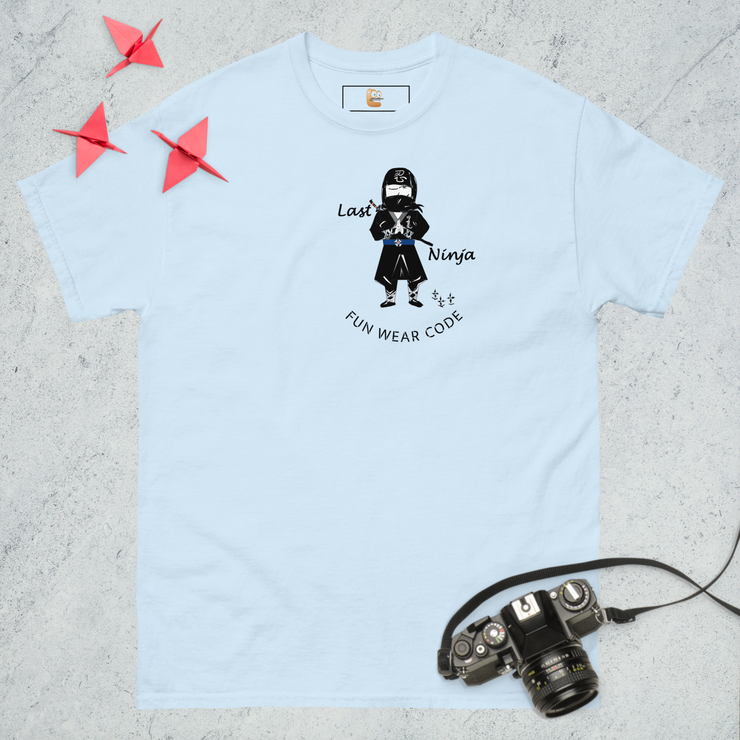 Light blue t-shirt with a playful "Last Ninja" graphic design and text "FUN WEAR CODE", placed on a textured surface with a camera and origami birds.