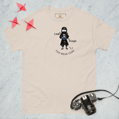 Beige t-shirt with a playful "Last Ninja" graphic design and text "FUN WEAR CODE", placed on a textured surface with a camera and origami stars.